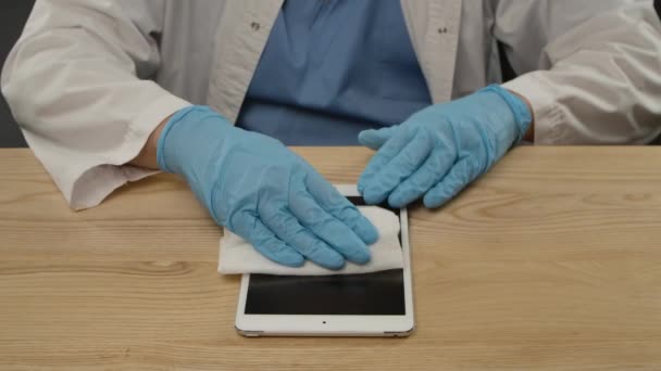 Ein Mann sitzt in einem medizinischen Schutzanzug an einem Schreibtisch und behandelt einen Notizblock mit einem Antiseptikum und einer Serviette. Hände in blauen Handschuhen schließen sich. Zeitlupe. — Stockvideo