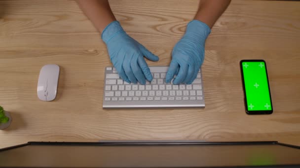 Vrouwen handen in beschermende blauwe handschoenen die typen op het toetsenbord in het kantoor of thuis. Smartphone met groen scherm en chroma toets met verticale schermpositie. Handen dicht. Langzame beweging. — Stockvideo