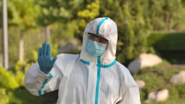 Koruyucu takım elbiseli, gözlüklü ve maskeli bir gencin portresi kameraya bakar ve jestlerin durduğunu gösterir, tamam, el sallayın ve buraya gelin. Ağır çekim. Kapat..