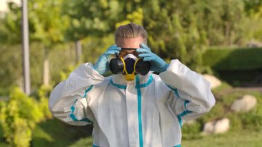 Koruyucu takım elbiseli ve eldivenli genç bir adamın portresi kameraya bakar ve bir solunum cihazı ve güvenlik gözlüklerinden kurtulur. Corona virüsünden korunma kavramı. Ağır çekim. Kapat..