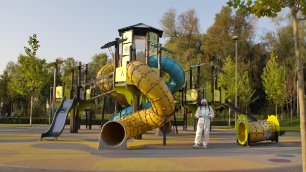 Um trabalhador em um respirador e traje de proteção descontamina passeios infantis e desliza em um parque público. Um homem desinfecta as superfícies dos playgrounds em quarentena do covid-19. Movimento lento. — Vídeo de Stock