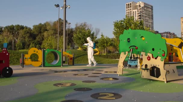 Un operatore sanitario in tuta bianca protettiva, respiratore, occhiali e guanti disinfetta un parco giochi con scivoli e attrazioni all'aperto in una giornata di sole. Rallentatore. — Video Stock