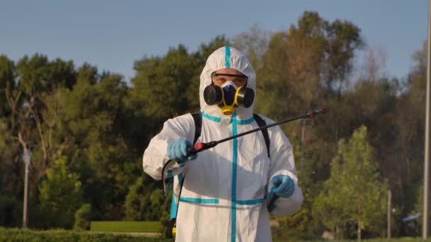 Un operaio disinfetta all'aperto in un parco pubblico con uno spray di disinfettante antibatterico. Un uomo con una maschera antigas respiratoria e una tuta protettiva con una rondella a pressione da vicino. Rallentatore. — Video Stock