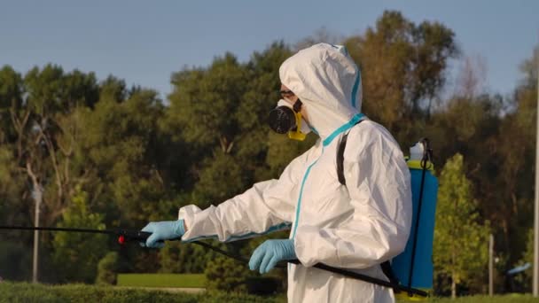 Vista lateral de um trabalhador desinfetando ao ar livre em um parque público com um spray de desinfetante antibacteriano. Homem com máscara de gás respiratório e um fato de proteção com uma arruela de pressão de perto. Movimento lento. — Vídeo de Stock