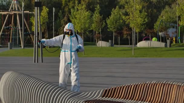 Un homme en combinaison de protection désinfecte les bancs d'un parc public avec des produits chimiques pour prévenir la propagation du coronavirus, une pandémie dans une ville en quarantaine. COVID-19. Mouvement lent. — Video