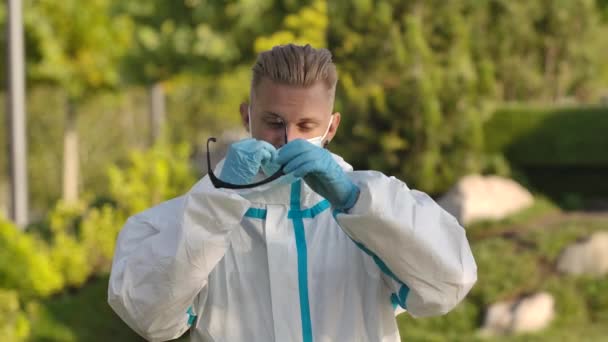 Porträtt av en ung man i vit skyddsdräkt tittar på kameran och tar av sig skyddsglasögon och en medicinsk mask. Hälsoskydd mot koronavirus. Sakta i backarna. Närbild. — Stockvideo