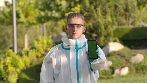 Portret van een jongeman in een beschermend pak, bril en handschoenen kijkt naar de camera en heeft een smartphone met groen scherm en chroma toets met verticale schermpositie. Langzame beweging. Sluiten.. — Stockvideo