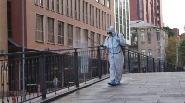 Beyaz koruyucu takım elbiseli bir hademe ve solunum cihazı viral enfeksiyonlara karşı basınç dezenfektanı ile şehir sokaklarının kaldırımlarını ve tırabzanlarını temizliyor. Dezenfekte. Karantinaya alın. Yavaş çekim