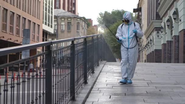Een werkende man in een beschermend pak en een masker desinfecteert, spuit chemicaliën op het oppervlak van de reling van het coronavirus. Sanitaire maatregelen op openbare plaatsen tijdens quarantaine. Langzame beweging. — Stockvideo
