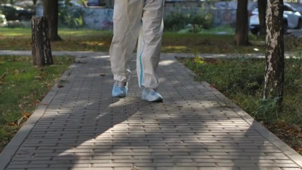 Bottom view of a virologist in a white protective suit disinfects paving slabs in a public park. The man uses a high pressure antiseptic solution. COVID-19. Cleaning concept. Slow motion. — Stock Video