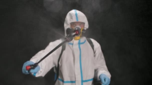 Portret van een man in een beschermend pak met een hogedrukapparaat desinfecteert met een antiseptische spray op een zwarte achtergrond in de studio. Langzame beweging. Sluiten.. — Stockvideo