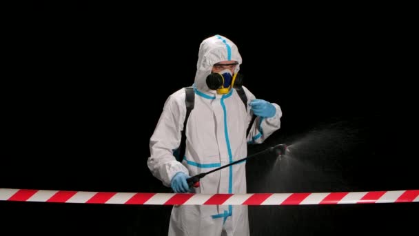 The view from behind the signal tape a man in a protective uniform and a respirator walks and disinfects the area on a black background in the studio. Coronavirus prevention concept. Slow motion — Stock Video