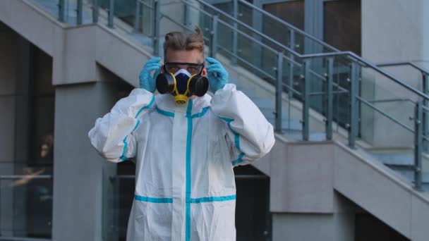 Portrait of a young man in a protective suit, respirator and gloves looks into the camera, puts on safety glasses and then takes off his glasses and respirator. Slow motion. Close up. — Stock Video