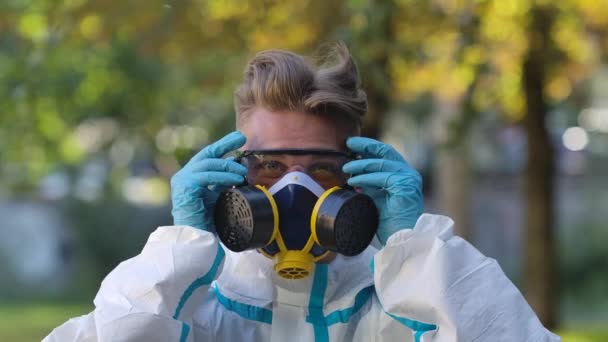 Porträtt av en ung man i skyddsdräkt, respirator och handskar tittar in i kameran, sätter på sig skyddsglasögon och tar sedan av sig glasögon och respirator. Sakta i backarna. Närbild. — Stockvideo