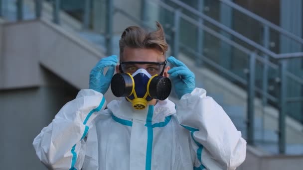 Porträtt av en ung man i skyddsdräkt, respirator och handskar tittar in i kameran, sätter på sig skyddsglasögon och tar sedan av sig glasögon och respirator. Sakta i backarna. Närbild. — Stockvideo