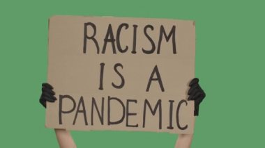 Siyah eldivenli bilinmeyen birinin elleri karton bir poster kaldırıyor. RACISM bir pandemiktir. Eşitlik ve birlik kavramı. Yeşil ekran, krom anahtar. Kapatın. Yavaş çekim.
