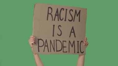 Kadın eller, karton bir kutudan üzerinde RACISM bir pandemiktir yazan bir poster tutuyorlar. Irkçılığa ve polis zulmüne karşı protesto. Yeşil ekran, krom anahtar. Kapatın. Yavaş çekim.