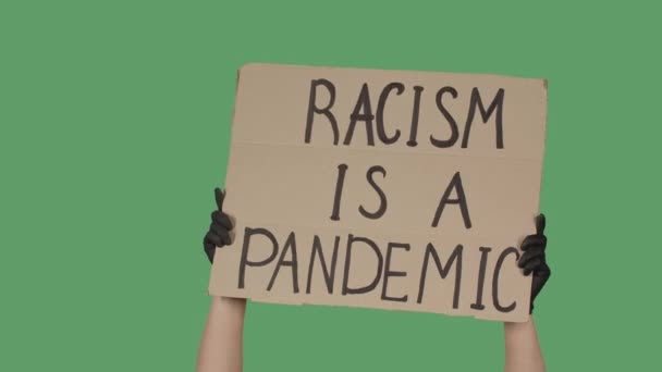 Kvinnliga händer i svarta handskar plockar upp en affisch från en pappkartong som läser RACISM IS A PANDEMIC. Kämpa för jämlikhet och enhet. Isolerade en grön skärm, kromnyckel. Närbild. Långsamma rörelser. — Stockvideo