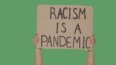 Elleri, üzerinde RACISM yazan bir posteri yeşil ekranın arka planında bir PANDEMIC 'tir, kroma tuşlu. Irkçılık yapma, ırkçılık yapma. Kapatın. Yavaş çekim.