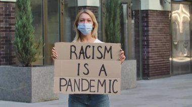 Tıbbi maskeli genç bir kadın, halka açık bir yerde, elinde bir karton poster olan bir RACISM ile duruyor. Toplumda ırkçılık, hoşgörü ve politik doğruluk yok. Ağır çekim. Kapat..