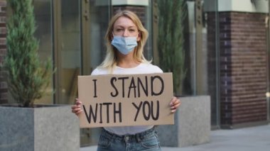 Tıbbi maske takan beyaz kadın protestocu seninle durduğum sloganın yazılı olduğu bir karton poster tutuyor. Irkçılık kavramını bırak. Ağır çekim. Açık hava portresini kapat..
