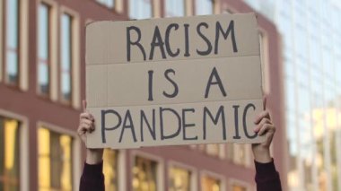 Erkek eller, üzerinde RACISM yazan bir posteri şehrin arka planına karşı bir pandemiktir. Irkçılık yapma, ırkçılık yapma. Kapatın. Yavaş çekim.