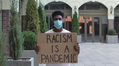 Tıbbi maskeli genç bir Afro-Amerikalı elinde bir karton posterle açık havada bir pandemikte duruyor. Sokak gösterisi. Ağır çekim. Kapat..