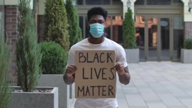 Siyah Yaşamlar Önemli yazılı bir poster tutan tıbbi maskeli Afro-Amerikan bir adam. Beyaz tişörtlü siyah öğrenci bir şehir caddesinde duruyor. Sokak gösterisi. Ağır çekim. Kapat..