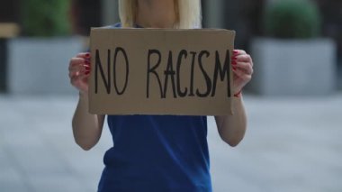 Beyaz kadın protestocuların elindeki karton posterde RACISM yok. Poster ve elin yakın çekimi. Irkçılık ve polis zulmüne karşı dayanışma içinde. Şehir sokak protestosu. Yavaş çekim.