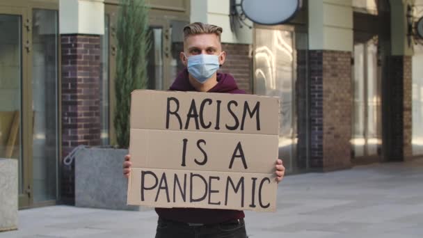 Młody człowiek w masce medycznej stoi z tekturowym plakatem RACISM IS A PANDEMIC in a public place outdoor. Brak rasizmu, tolerancji i poprawności politycznej w społeczeństwie. Zwolnij trochę. Zamknij się.. — Wideo stockowe