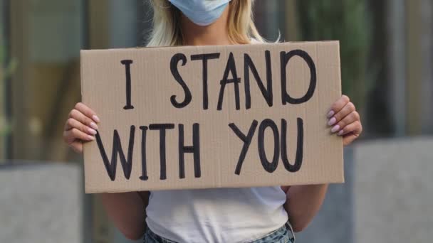 저는 의료용 마스크를 쓴 여성 시위 운동가의 손에 판지로 된 포스터에 여러분과 함께 서 있습니다. 포스터와 손을 클로즈업 합니다. 격리 기간 동안 도시의 거리 시위. 느린 동작. — 비디오