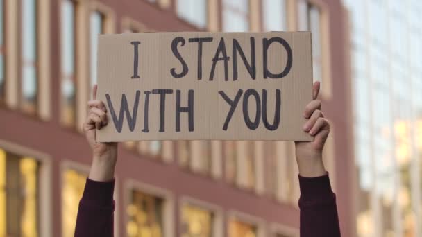在城市的背景下，男性的手拿着一张标语牌，上面写着"我和你站在一起" 。抗议海报在室外被贴上.街头示威。靠近点慢动作. — 图库视频影像