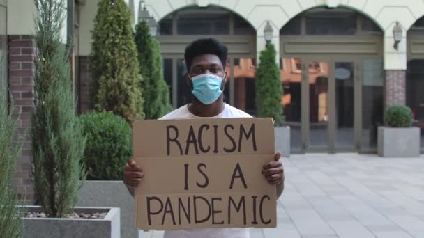 医療用マスクの若いアフリカ系アメリカ人男性が段ボールのポスターRACISMは公共の屋外の場所でパンデミックです。ストリートデモンストレーション。スローモーション。閉じろ!. — ストック動画