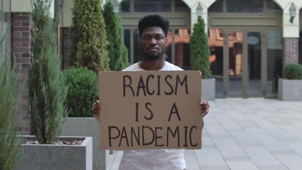 若いアフリカ系アメリカ人の男性が段ボールのポスターRACISMは、公共の屋外の場所でパンデミックです。ストリートデモンストレーション。スローモーション。閉じろ!. — ストック動画