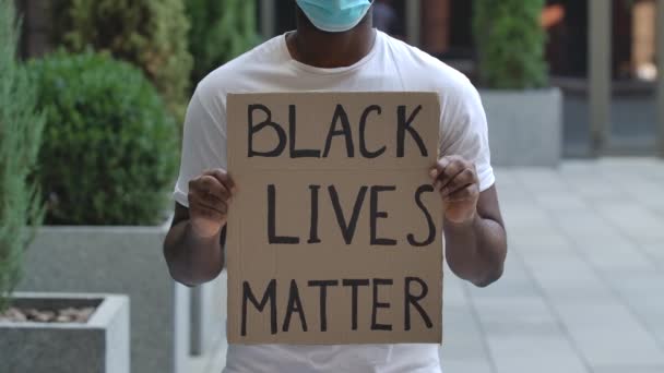 Um afro-americano está no meio da rua segurando um cartaz de papelão na frente dele que lê Black Lives Matter. Movimento lento. Feche o cartaz e o tronco de um homem. — Vídeo de Stock