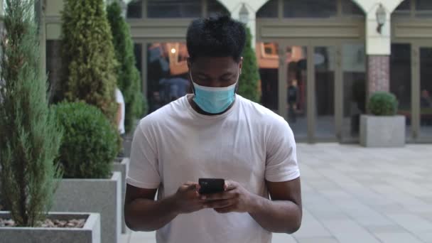 Porträtt av ung afroamerikansk man i skyddande medicinsk mask sms:ar på sin smartphone. Hälsoskydd för koronavirus. Sakta i backarna. Närbild. — Stockvideo