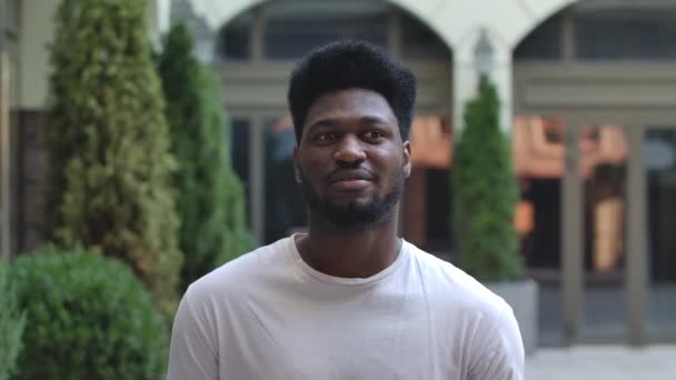 Portrait de jeune homme afro-américain regardant la caméra et dit quelque chose. Brunet élégant dans un T-shirt blanc sur le fond de la rue à l'extérieur. Au ralenti. Gros plan. — Video