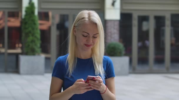 Retrato de una encantadora joven escribe un mensaje de texto en su teléfono inteligente se regocija y sonríe. Rubia en una blusa azul al aire libre en el fondo de la calle. En cámara lenta. De cerca.. — Vídeo de stock
