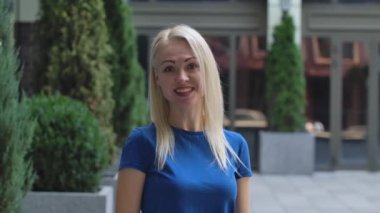 Çekici bir genç kadının portresi kameraya bakıyor ve neşeli ve gülümseyen biriyle konuşuyor. Caddenin arka planında mavi bluzlu bir sarışın var. Ağır çekim. Kapat..