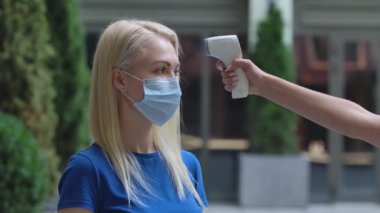 Koruyucu tıbbi maske takan ve sıcaklığı temas olmayan termometreyle ölçülen çekici genç bir kadının portresi. Corona virüsü sağlık koruma konsepti. Yan görüş. Ağır çekim. Kapat..
