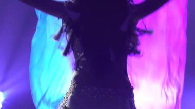 Beyaz doğulu bir kostüm giymiş, ipek kumaşla dans eden kadın dansçı. Duman ve mavi ışıklı karanlık bir stüdyoda çekildi. İnce, esnek bir vücudun siluetleri. Kapatın. Yavaş çekim.