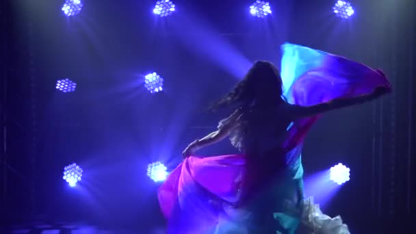 Danseuse du ventre dans un costume oriental blanc dansant avec un tissu de soie. Tourné dans un studio sombre avec de la fumée et un néon bleu. Silhouettes d'un corps souple mince. Ferme là. Mouvement lent. — Video