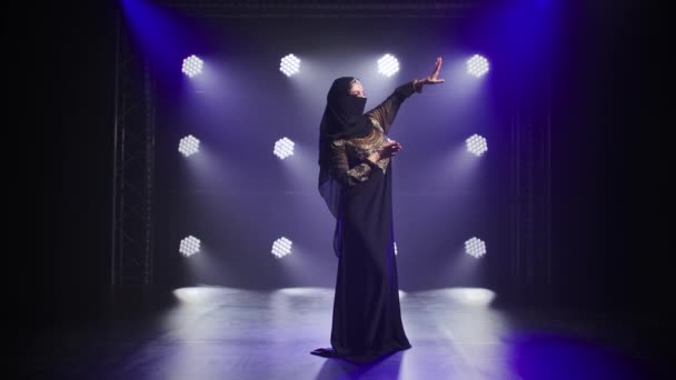 Beautiful belly dancer woman graceful dancing in a black oriental dress and hijab. Shot in a dark studio with smoke and neon lighting. Slow motion. — Stock Video