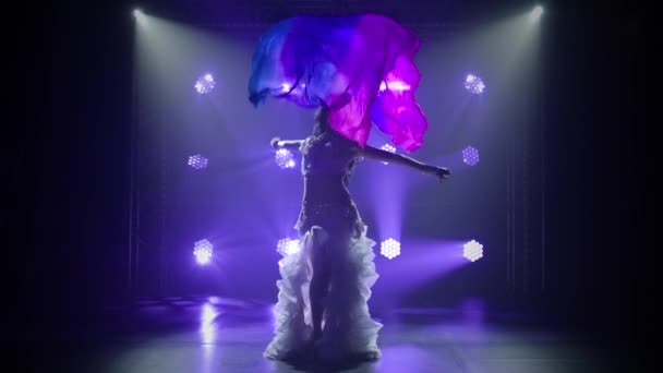 Female belly dancer in a white oriental costume dancing with silk cloth. Shot in a dark studio with smoke and blue neon lighting. Silhouettes of a slender flexible body. Slow motion. — Stock Video