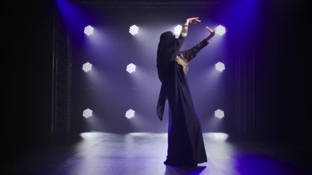 Belle danseuse du ventre femme gracieuse danse dans une robe orientale noire et hijab. Tourné dans un studio sombre avec de la fumée et un éclairage au néon. Mouvement lent. — Video