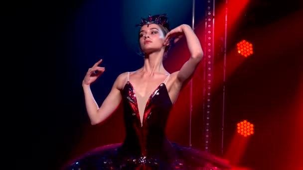 Graciös ballerina i en chic bild av en svart svan. Ung vacker flicka i svart tutu med röda paljetter och en krona. Skjuten i en mörk studio med rök och röd neonbelysning. Långsamma rörelser. — Stockvideo
