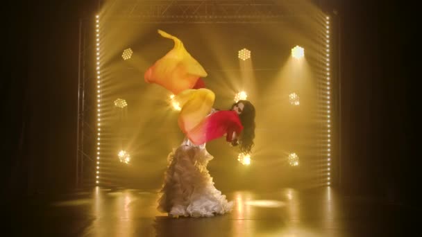 Танцовщица живота в белом восточном костюме танцует шелковые веерные ткани. Снято в темной студии с дымом и желтым неоновым освещением. Медленное движение. — стоковое видео