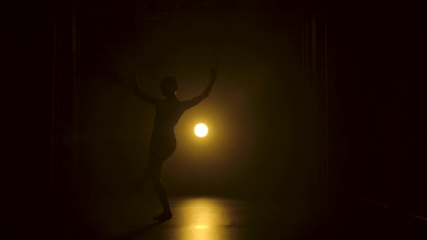 Silhueta uma bailarina flexível fina em bodysuit preto dança graciosa na escuridão sob um teatro holofotes amarelos de estúdio. — Vídeo de Stock