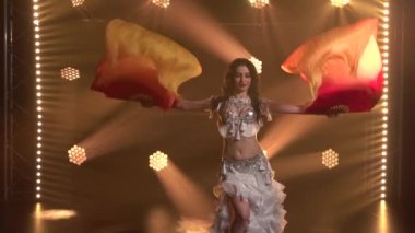 Beyaz doğulu kostümlü, ipek fan dokularıyla dans eden kadın dansçı. Duman ve neon ışıkla karanlık bir stüdyoda çekildi. Kapatın. Yavaş çekim.