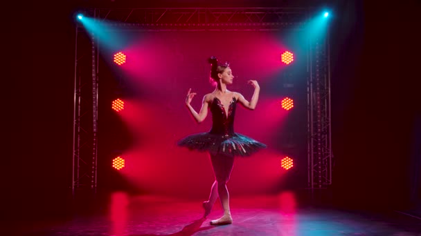 Půvabná baletka v elegantním obrazu černé labutě. Mladá krásná dívka v černé sukničce s červenými flitry a korunou. Natočeno v tmavém studiu s kouřem a červeným neonovým osvětlením. Zpomalený pohyb. — Stock video